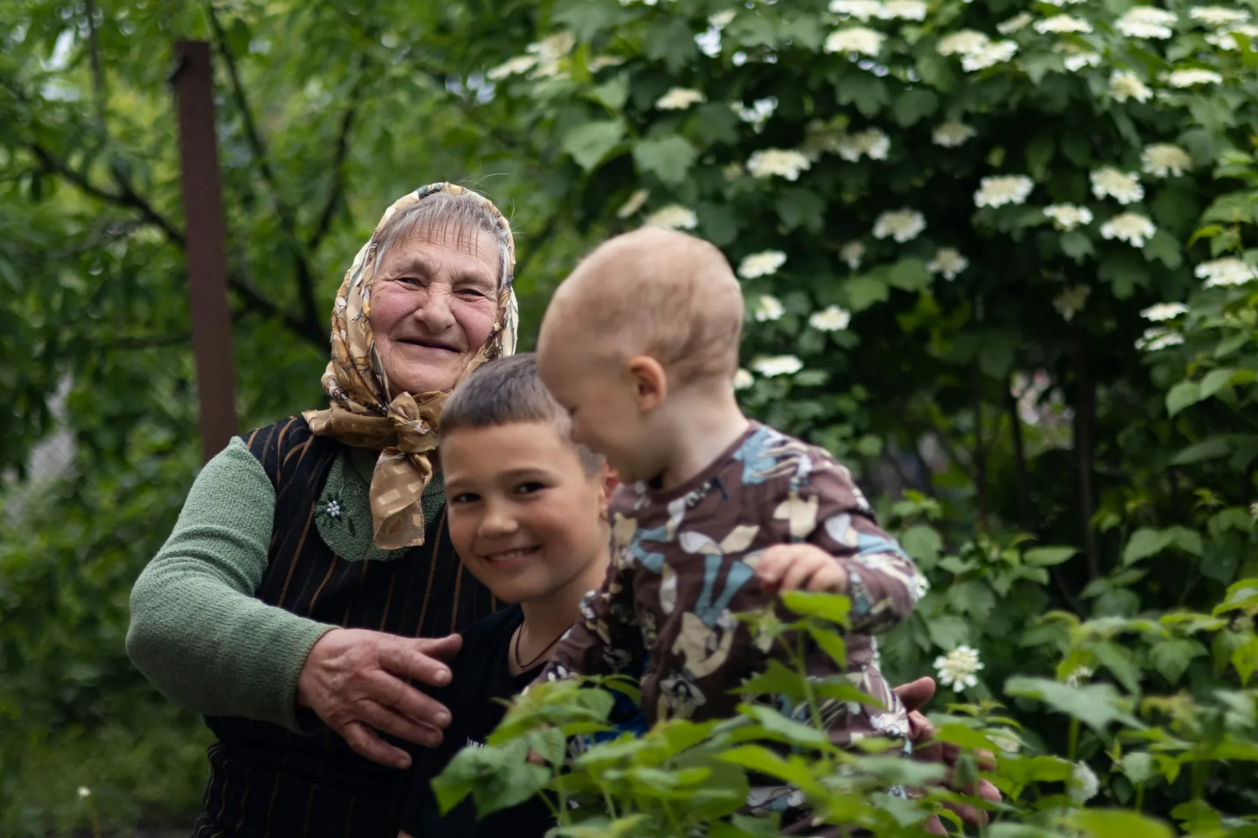 A smiling grandmother in a patterned headscarf with her arms around two young children in a leafy garden.