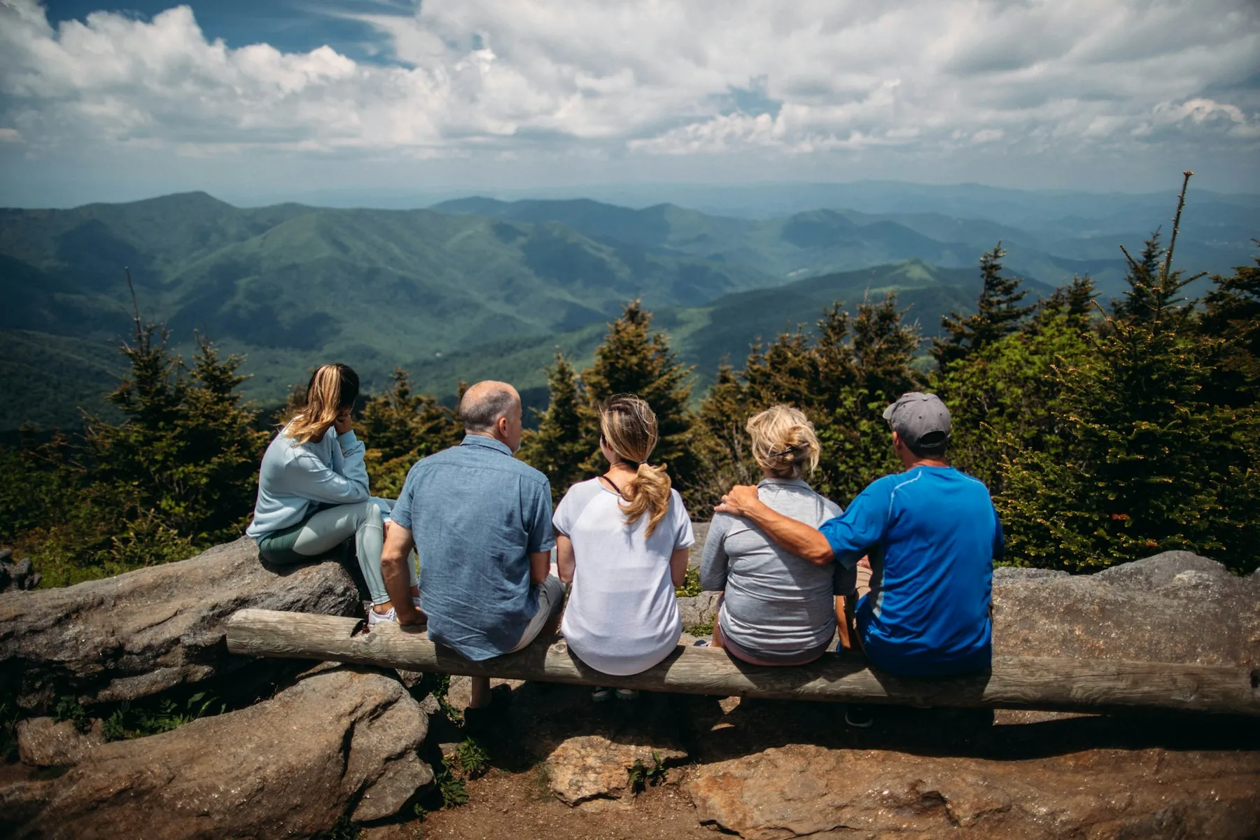 A family of five seen from behind, sitting together on a log bench and looking out over green mountain ridges.