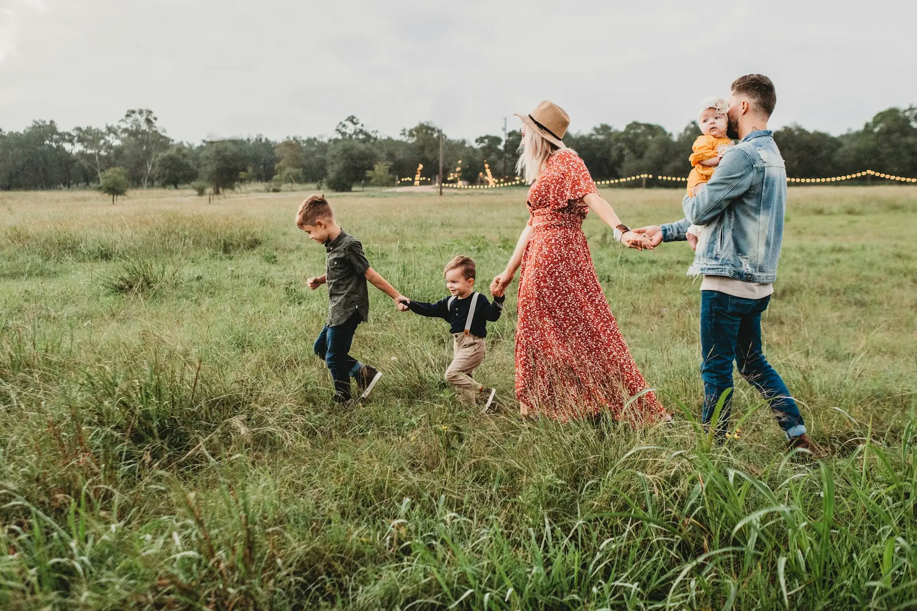 A family of five walking hand in hand in a line through tall grass at dusk, with string lights glowing at the edge of the field.