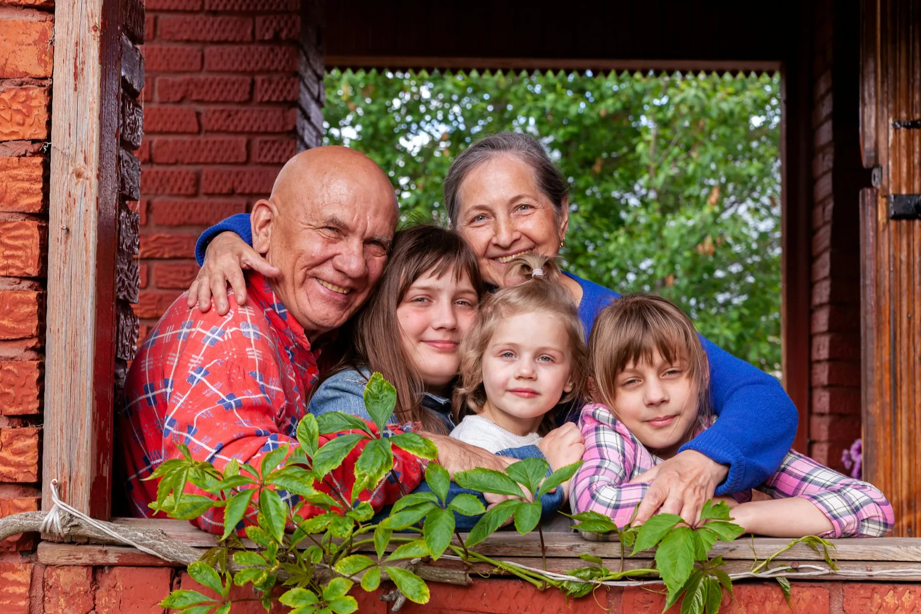 Grandparents hugging three grandchildren close at the open window of a red brick porch, with green leaves behind them.