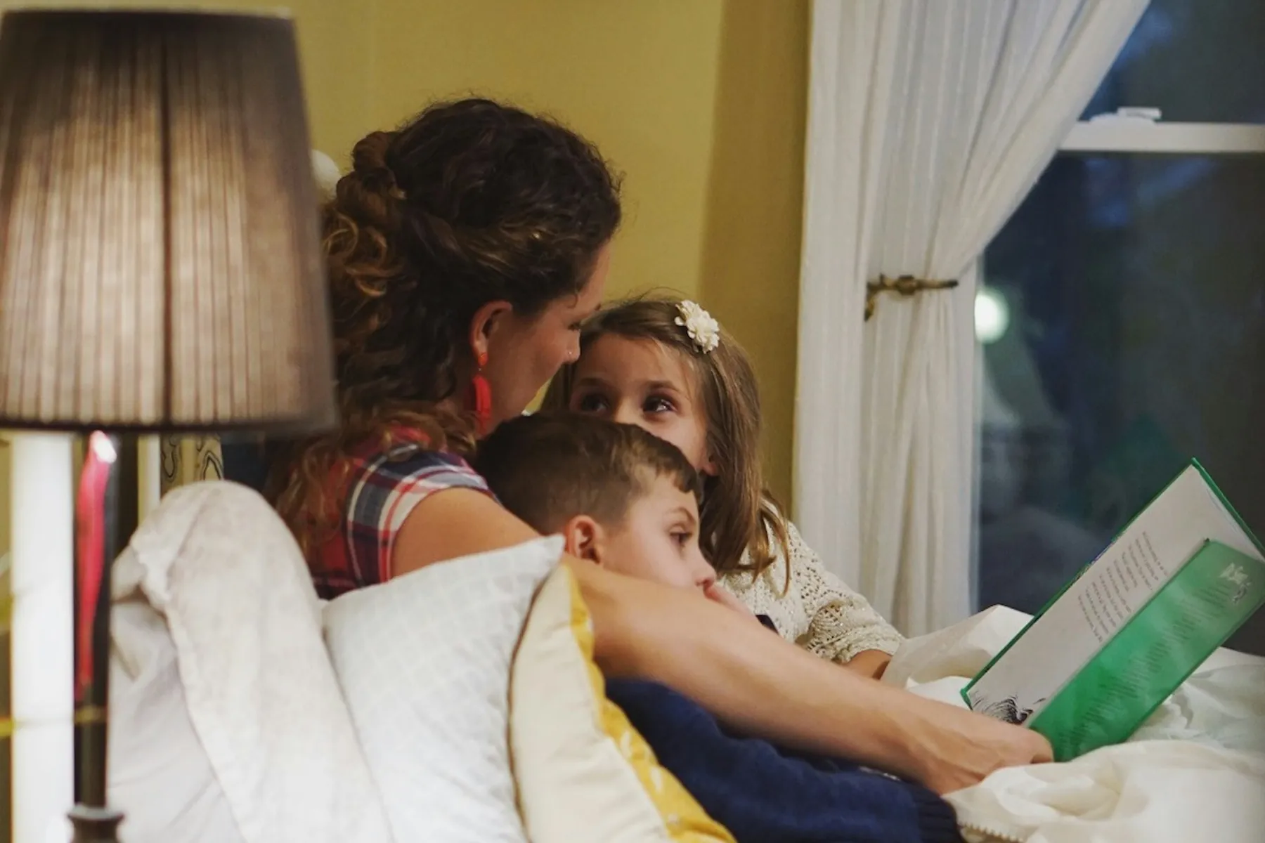A mother reading a picture book in bed with her son and daughter close beside her, a lamp and a window behind them.
