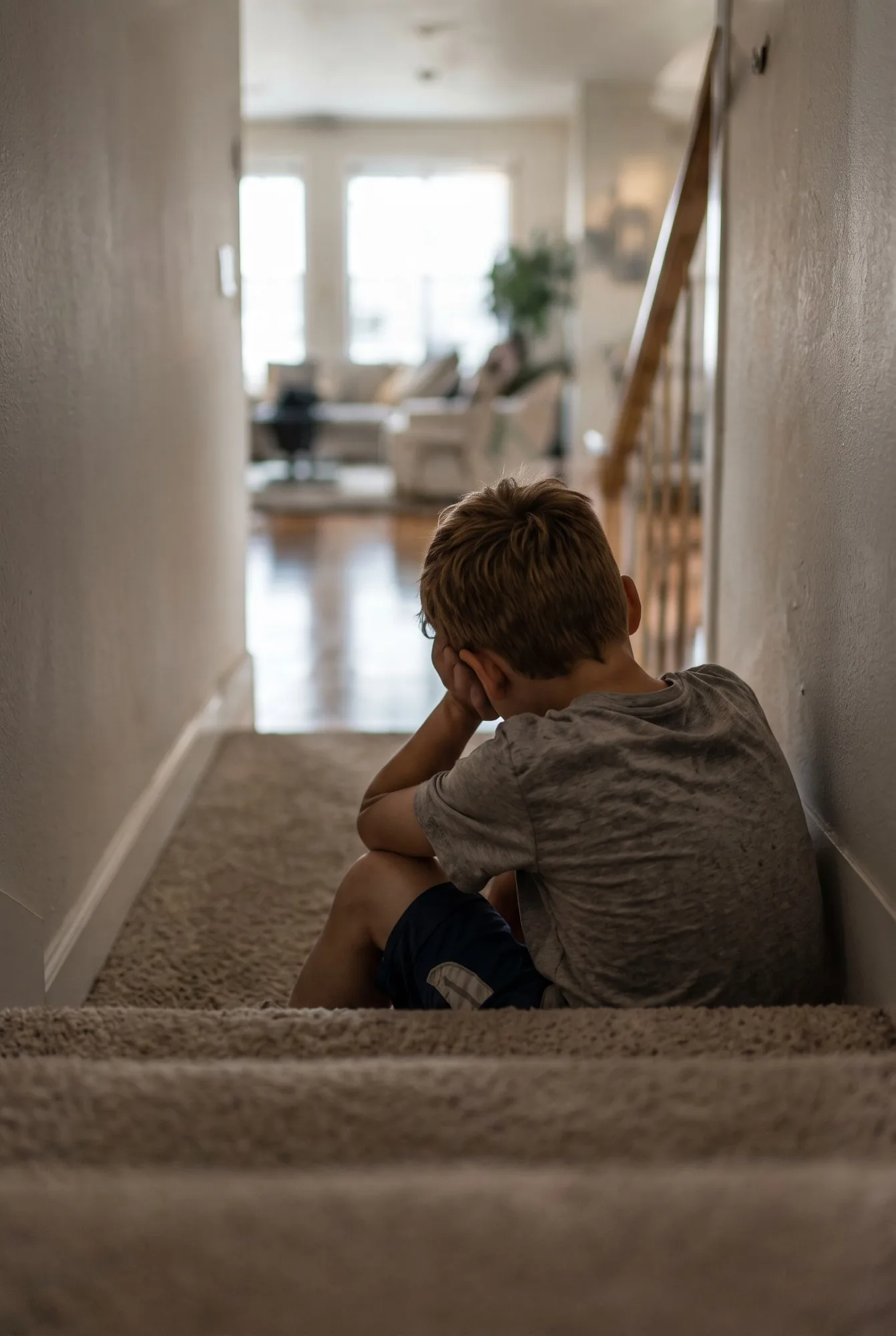 A young boy sits alone at the top of the stairs with his face in his hands, the bright living room far below.