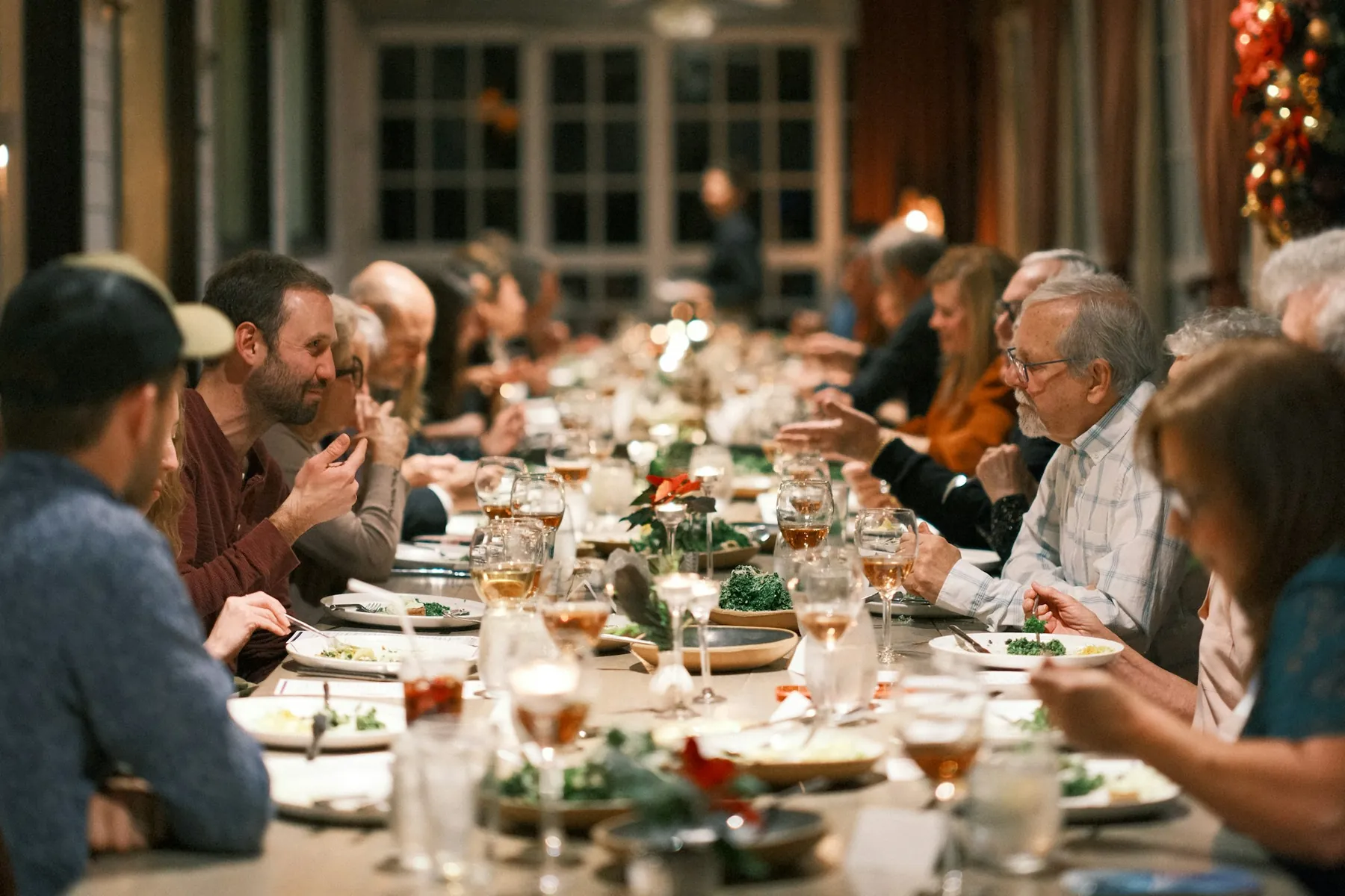 A long wooden table in a warm, lamplit room, with people of different ages deep in conversation over plates and candles.