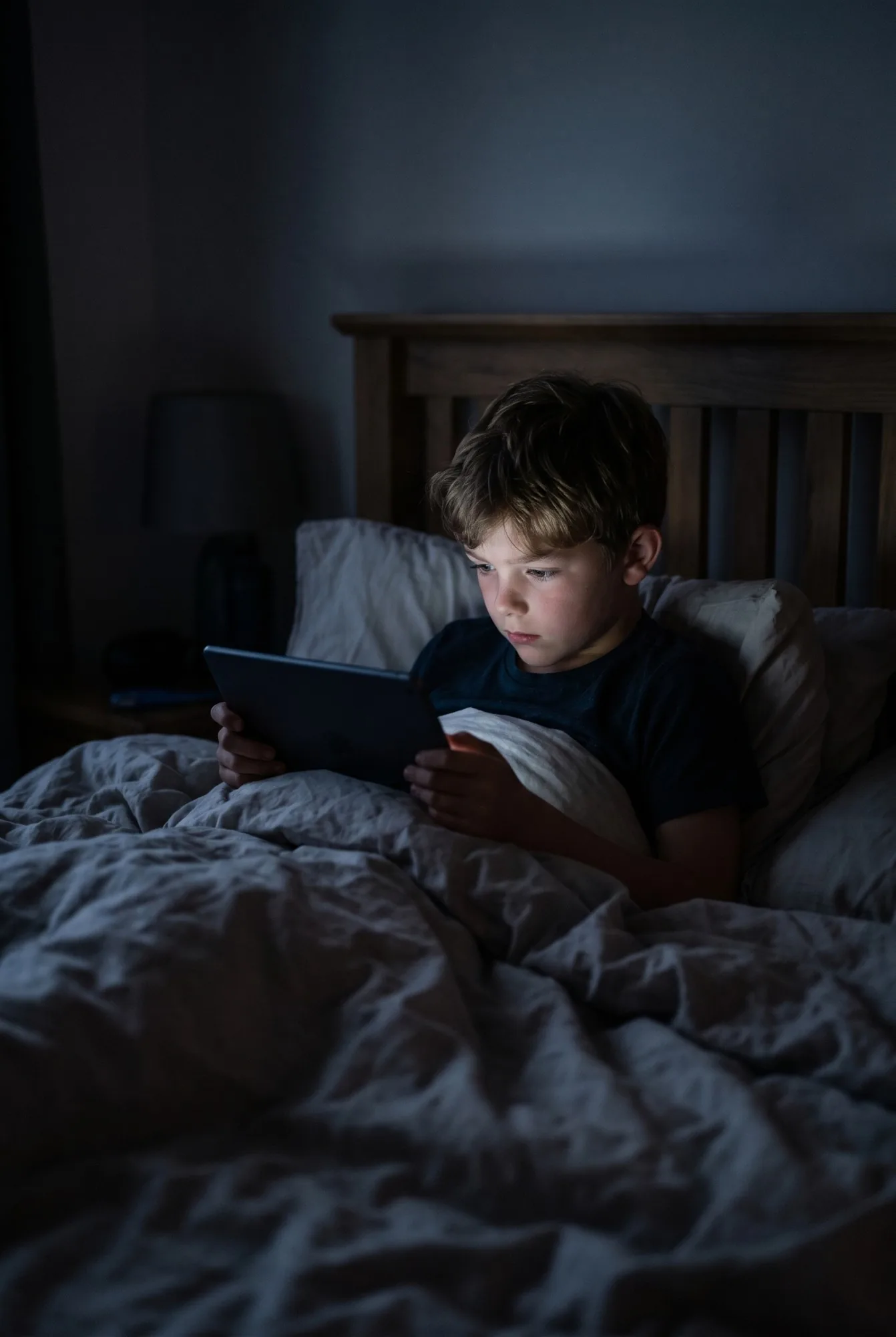 A young boy sits up in bed in a dark room, his face lit by a tablet.