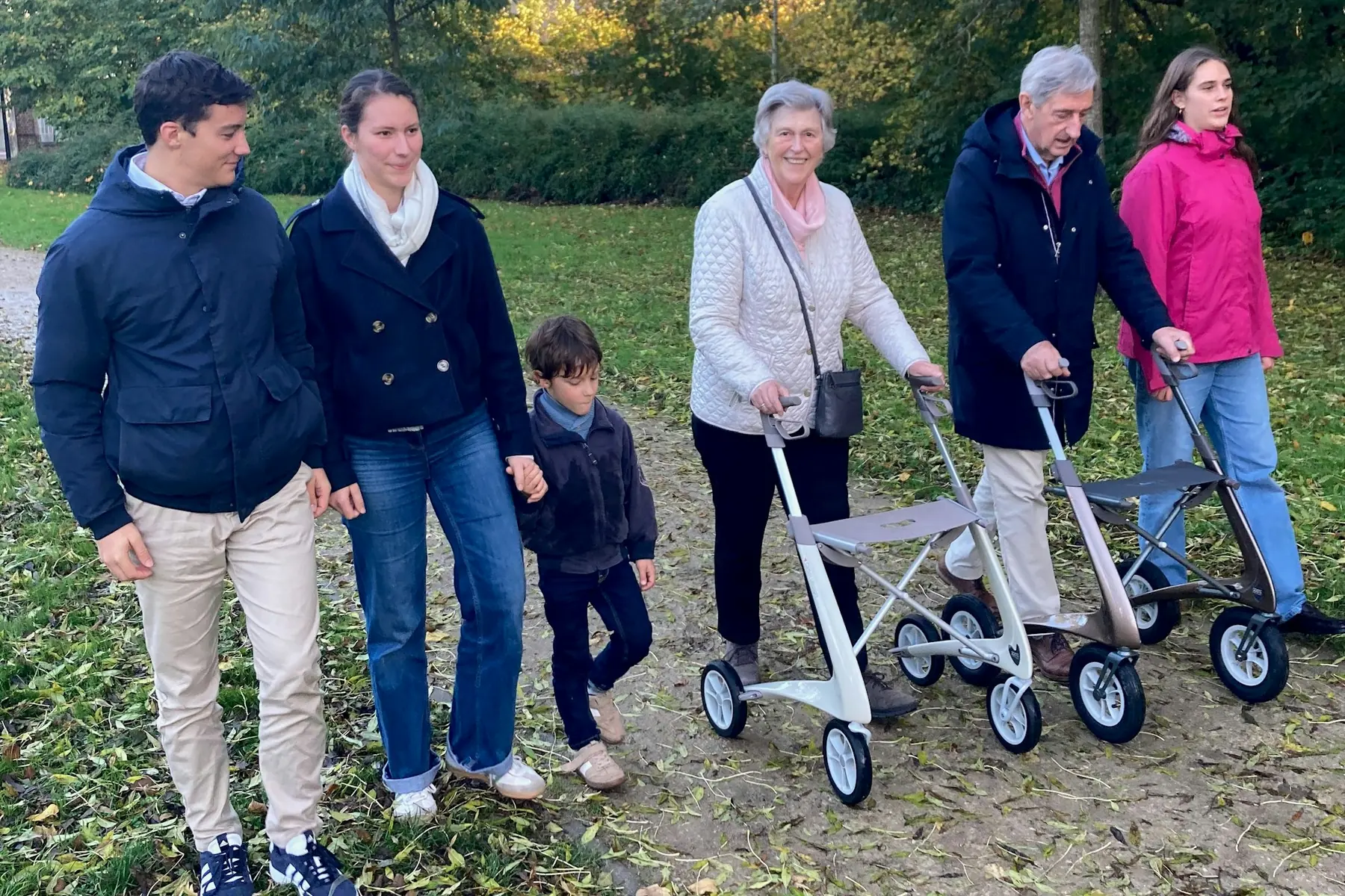 Three generations walking together on an autumn park path, with two grandparents using walkers and younger family beside them.