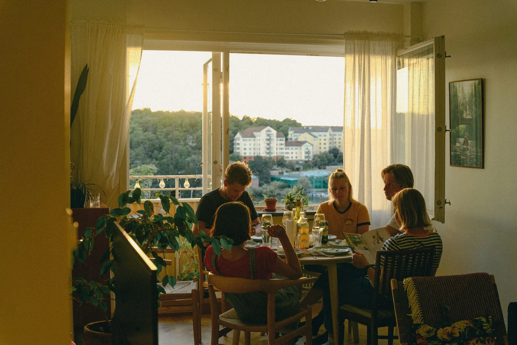 Five people sharing supper at a round table in front of an open balcony window in warm evening light, one reading from a picture book.